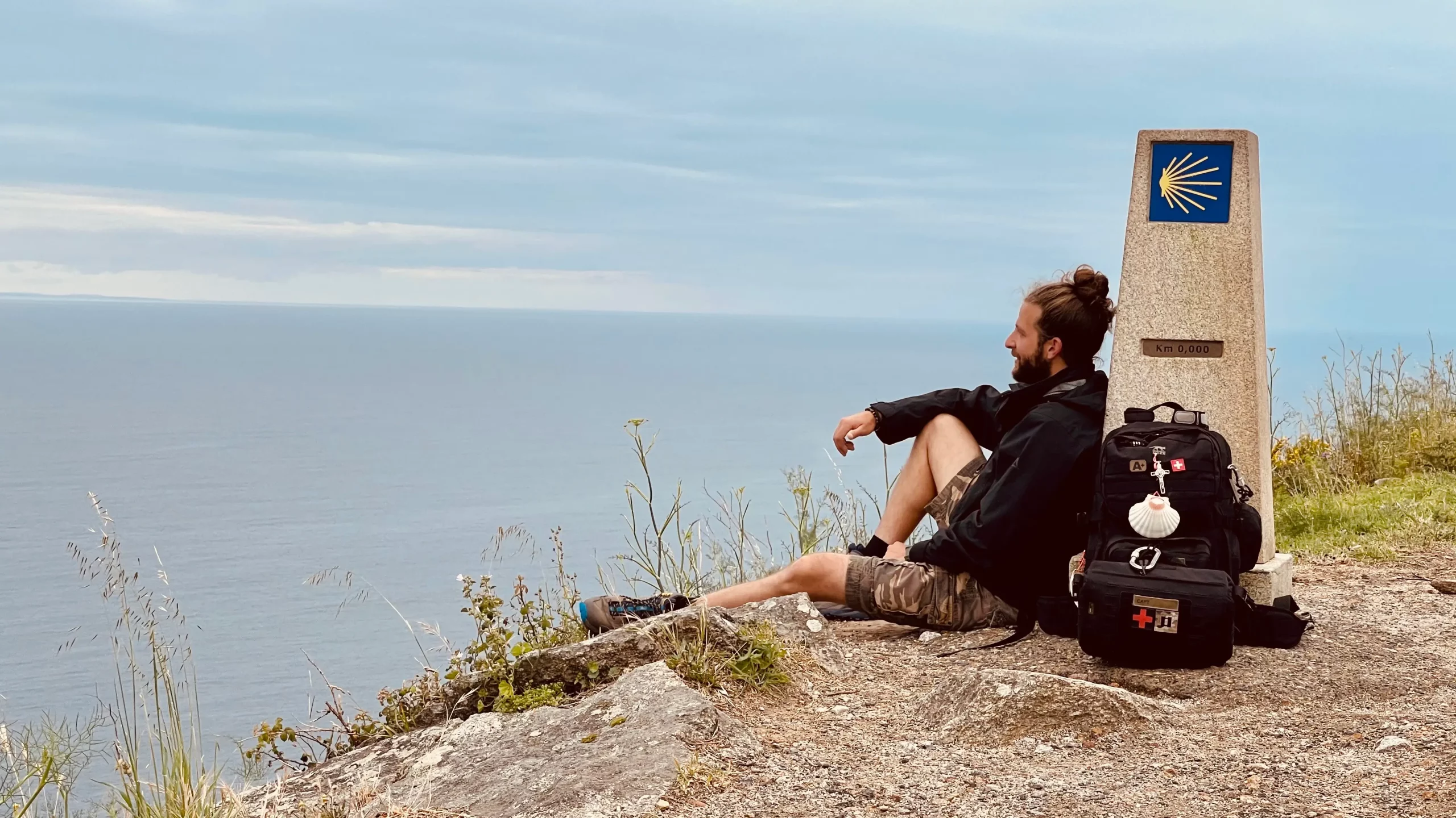 Fabio Sinicropi assis contre la borne du kilomètre 0 à Cabo Fisterra, son sac à dos à ses côtés, regardant l’horizon après son pèlerinage de Compostelle.