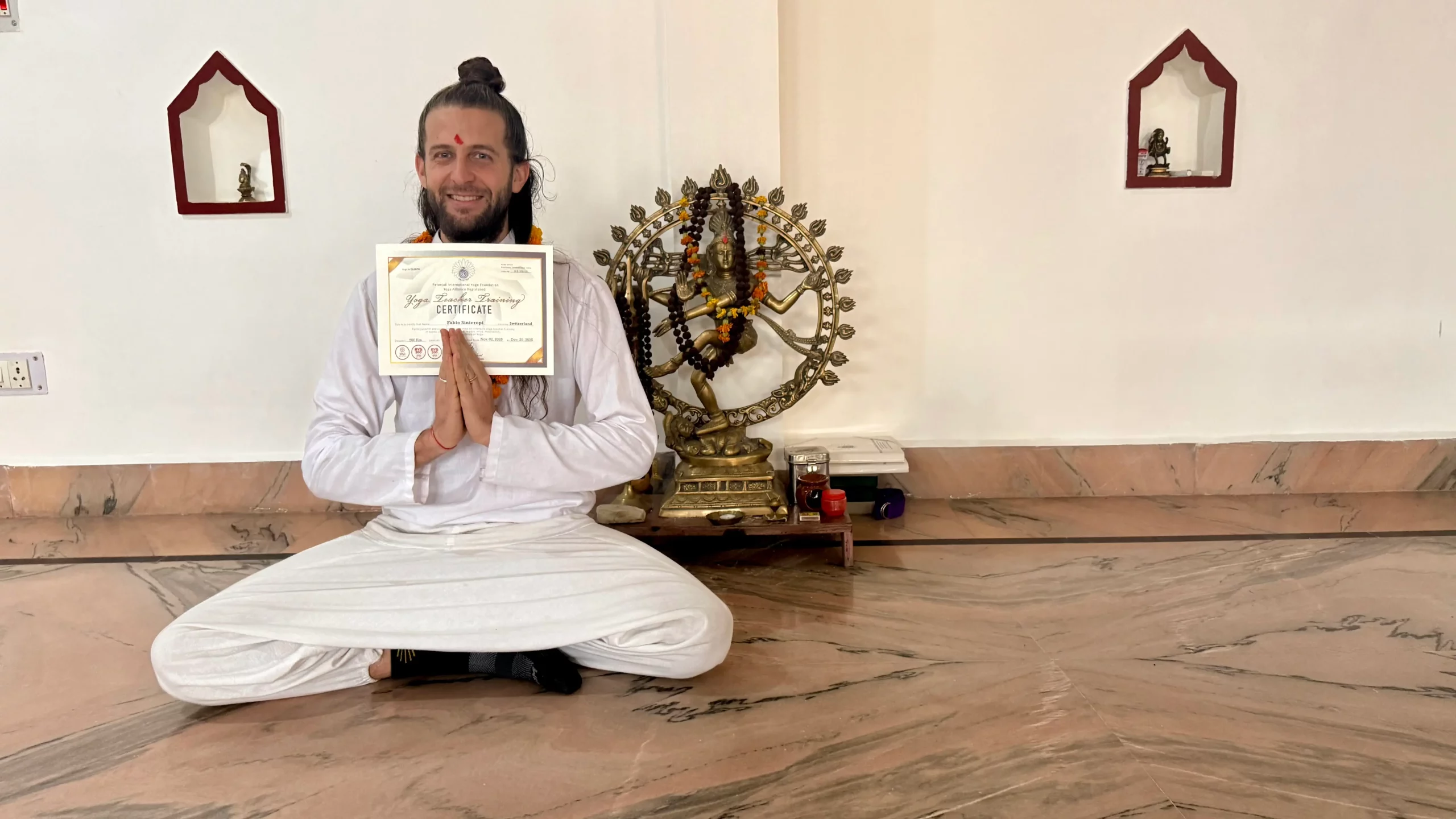 Fabio Sinicropi, souriant et vêtu de blanc, assis en tailleur pour présenter son certificat de formation de professeur de yoga devant une statue en bronze de Shiva, illustrant son apprentissage traditionnel en Inde.
