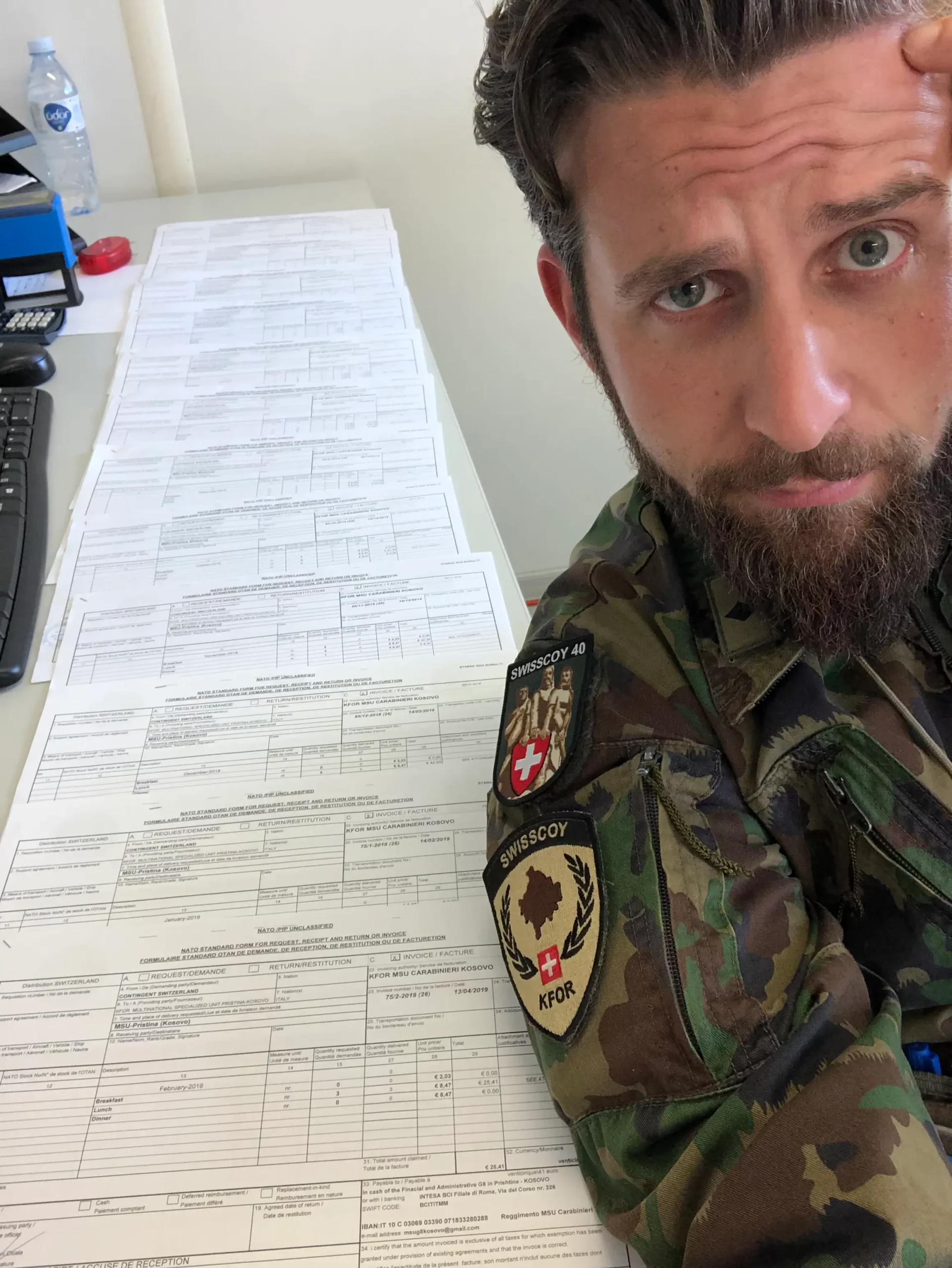 Fabio Sinicropi en uniforme militaire de la Swisscoy (KFOR) au Kosovo, l'air songeur devant une très longue file de documents administratifs sur un bureau, illustrant son ancienne carrière d'officier dans l'armée suisse.